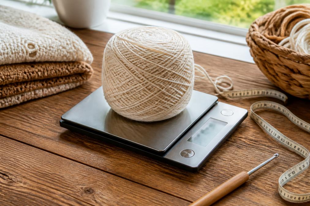Digital kitchen scale displaying yarn weight in grams, with a ball of yarn, measuring tape, and crochet hook arranged on a wooden crafting table with natural window lighting