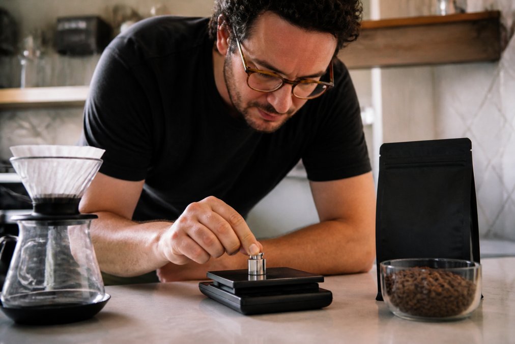 Close-up of hands placing a small calibration weight onto a digital coffee scale on a kitchen counter, with a bag of coffee beans and a pour-over dripper nearby