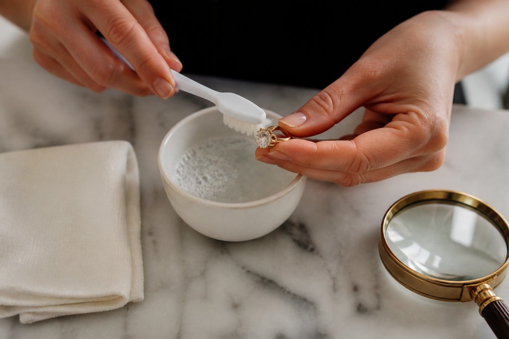 Close-up of hands gently cleaning an antique diamond ring with a soft-bristled toothbrush and mild soapy water in a small ceramic bowl, with a lint-free cloth and magnifying glass on a marble countertop