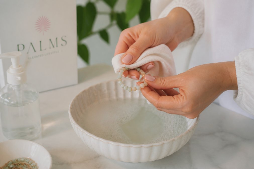 Close-up of hands gently cleaning a delicate beaded bracelet with a soft cloth and mild soap solution in warm water, showing careful technique and attention to detail