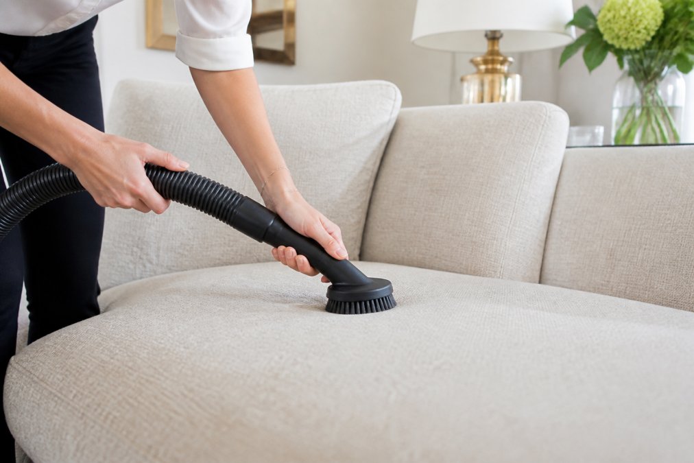 Hands using a soft brush vacuum attachment to gently clean a high-end fabric sofa in a bright living room.