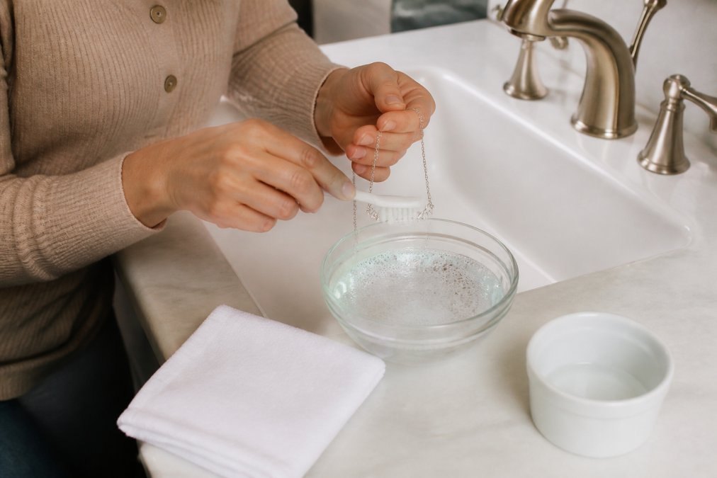 Person gently cleaning a personalized silver necklace with a soft brush and soapy water at a bathroom sink