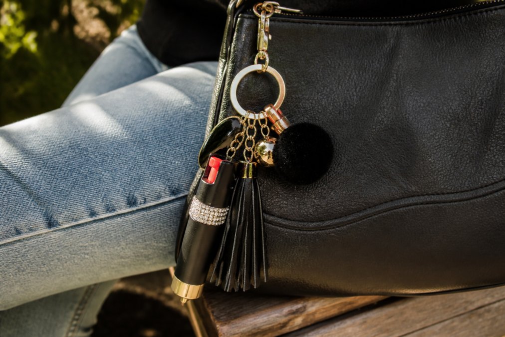 Close-up of a woman's keychain with stylish self-defense pepper gel attached to a designer leather purse, showing the compact gel canister alongside fashion accessories in natural afternoon sunlight