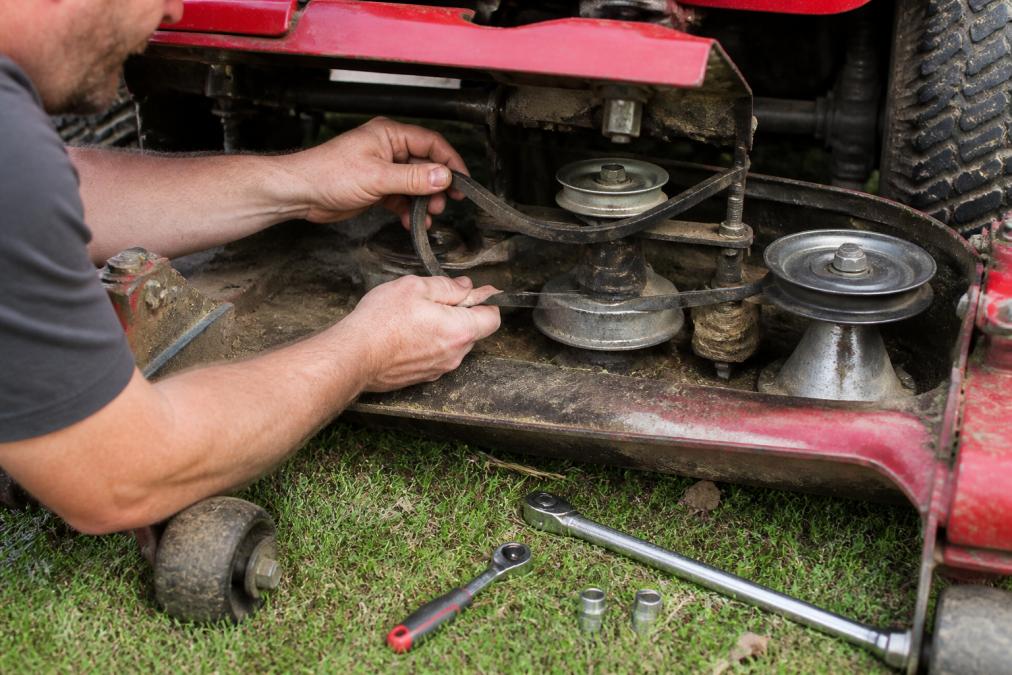 Technician's hands carefully examining the routing of a worn V-belt around pulleys and spindle assemblies on a riding mower deck, with the belt guide visible and tools positioned nearby on the grass