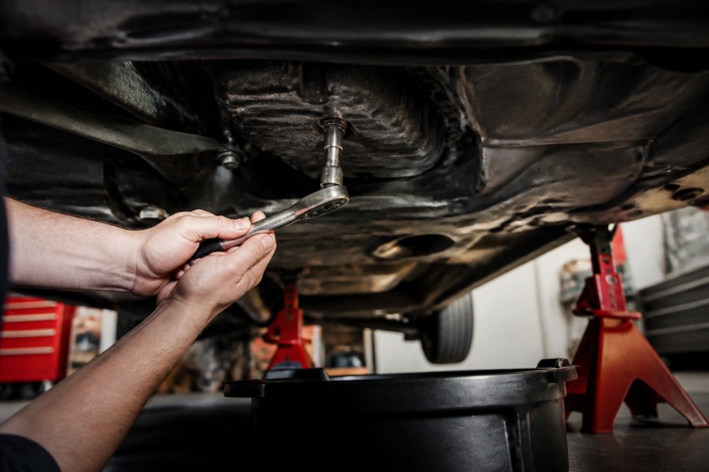Close-up of a mechanic's hands using a socket wrench to loosen the drain plug on a transmission pan, with a catch pan positioned underneath to collect fluid, car raised on jack stands in a home garage
