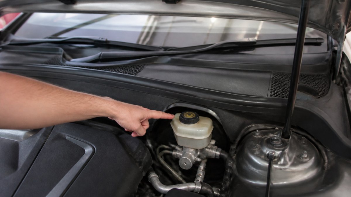 A person's hand pointing to the translucent brake fluid reservoir near the firewall in the engine bay of an Audi, with the hood raised and the reservoir cap clearly visible