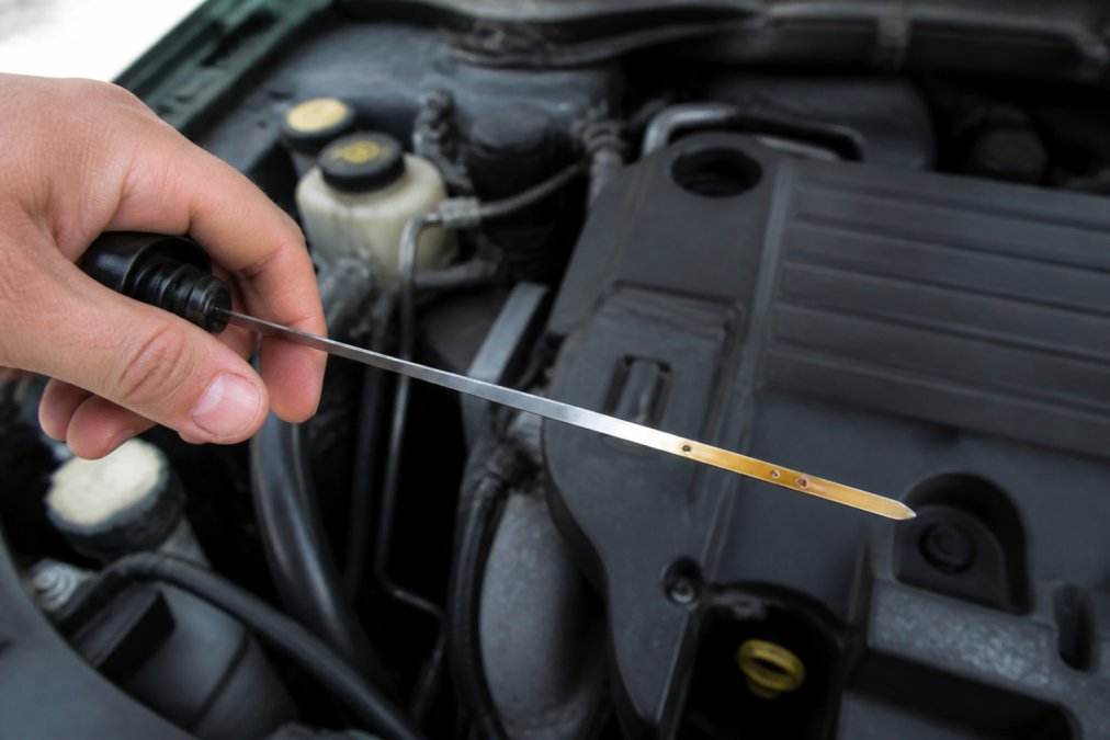 Close-up of hands pulling out an oil dipstick from under an open car hood, with the dipstick showing clear full and low level markings against the engine bay background