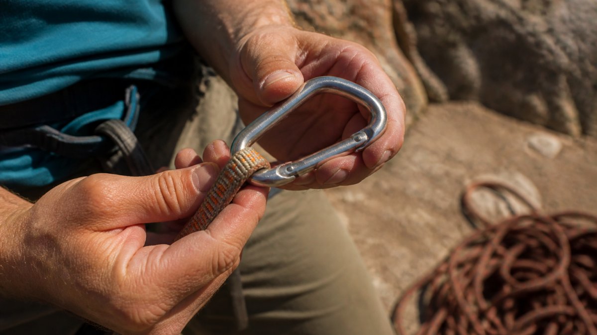 Close-up of a climber's hands holding a quickdraw with carabiner gate visible, examining it under natural sunlight with clear detail of the metal surface and sling attachment