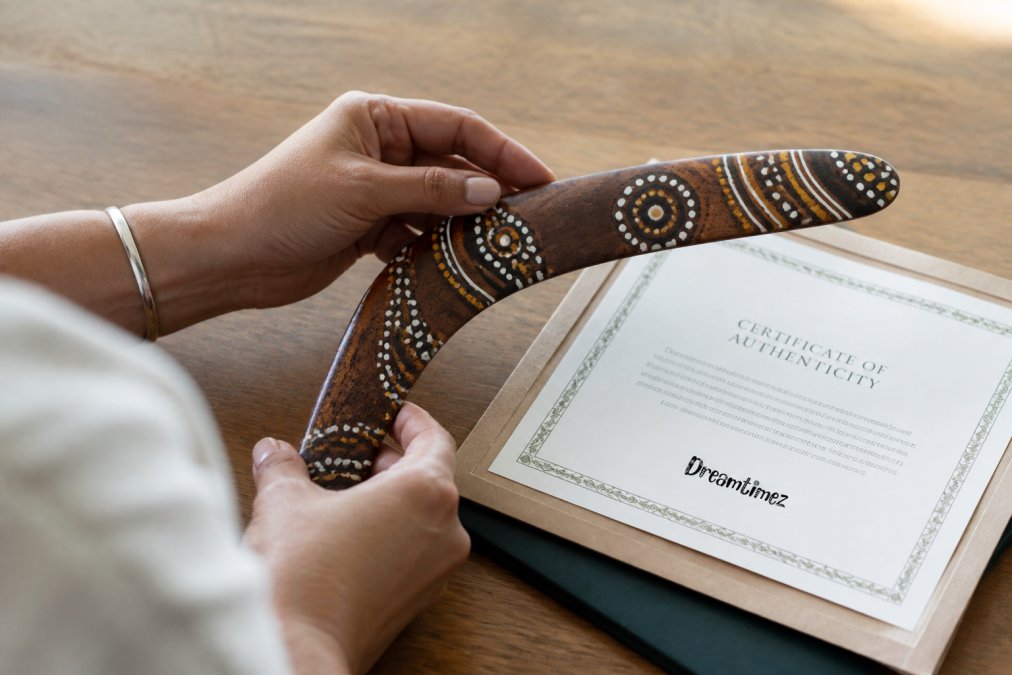 A close-up of hands holding a beautifully crafted item featuring Indigenous artwork, with the artist's certificate of authenticity visible nearby on a wooden table in soft natural light