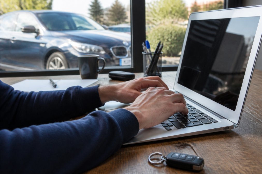 Close-up of hands typing a vehicle registration plate number into a laptop keyboard, with a used car visible through a window in the background in natural daylight