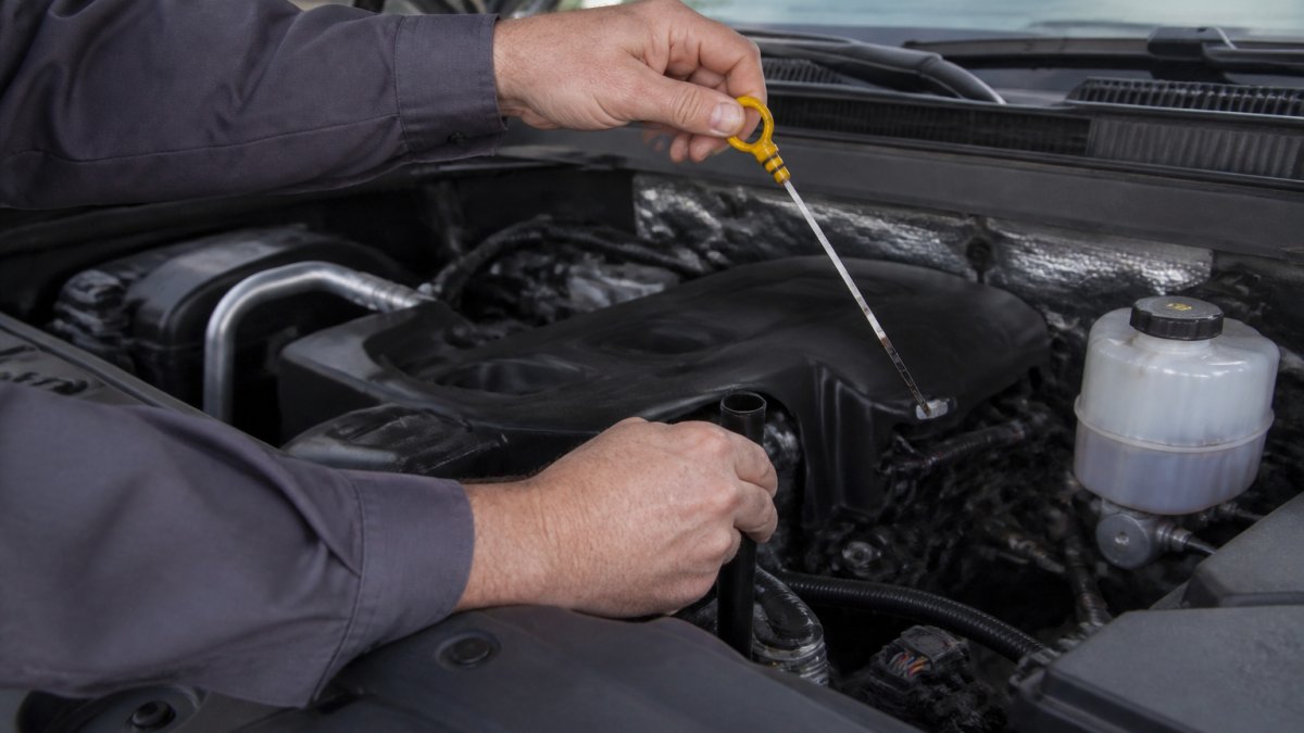 Professional technician's hands removing a transmission dipstick from under the hood of a GMC truck, showing the dipstick tube and handle clearly in natural daylight