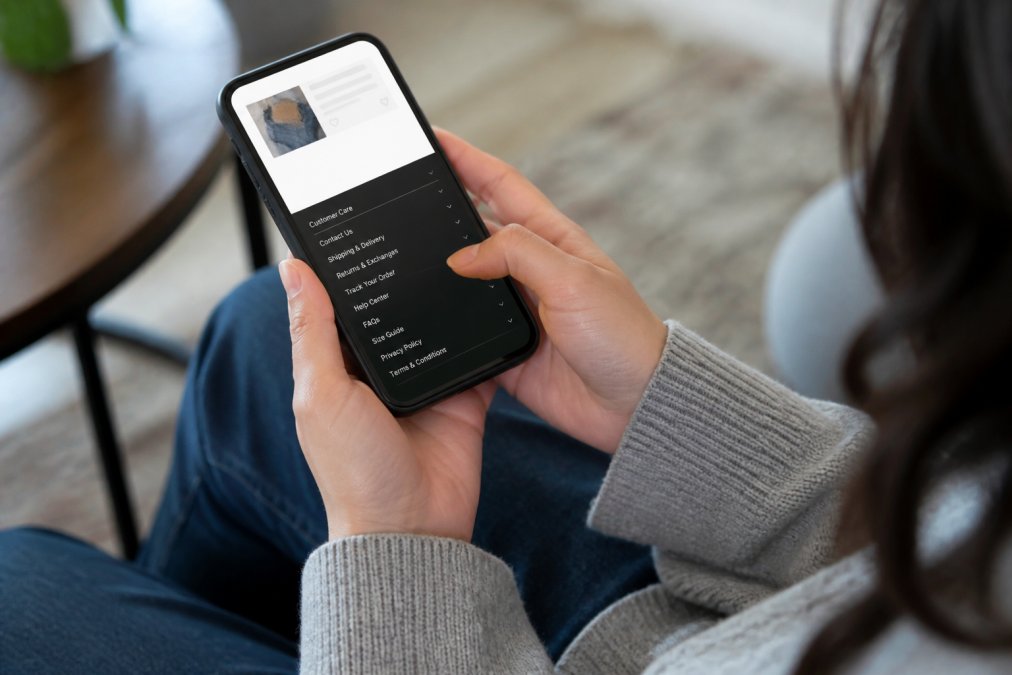 Woman's hands holding smartphone, scrolling through a brand's website footer showing return policy links, customer care options, and help center navigation with natural indoor lighting