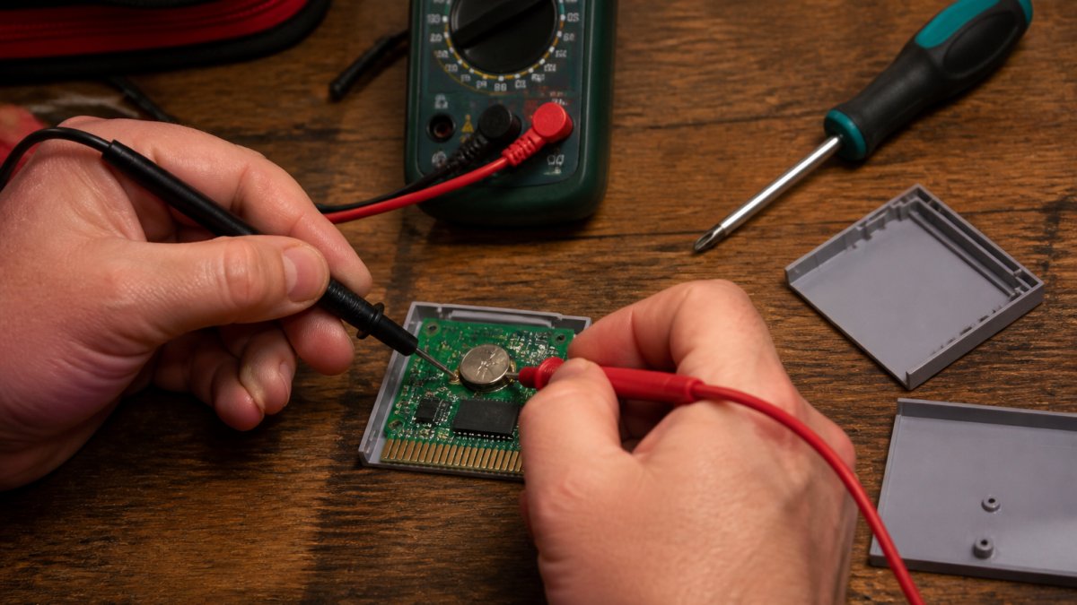 Hands using a multimeter to test voltage on retro game save batteries inside an opened Game Boy cartridge