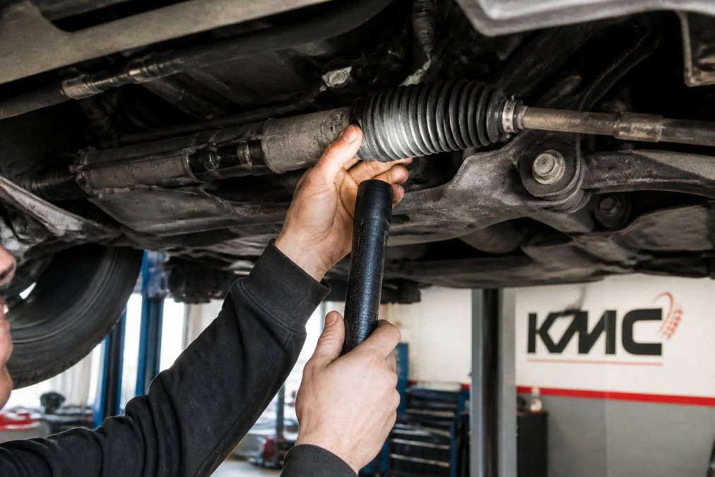 Close-up of a mechanic's hands using a torch to inspect a rubber steering rack boot on a car raised on a lift in a clean garage, showing the gaiter and clips clearly
