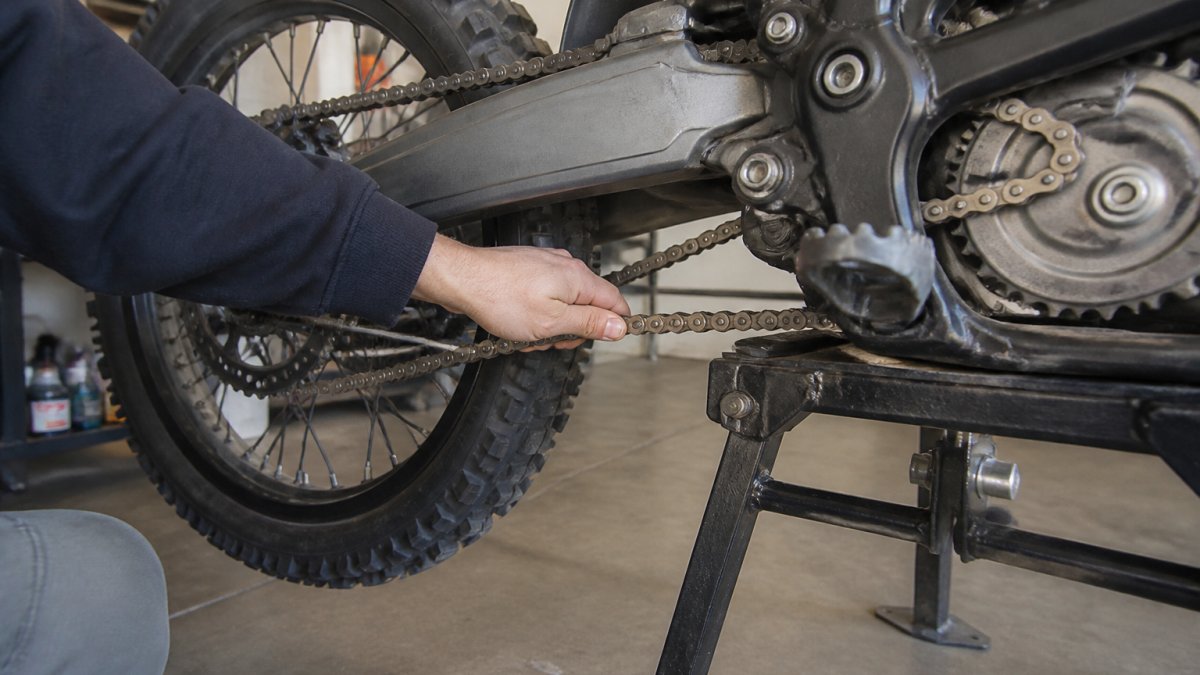 Close-up of a rider's hand pressing down on the lower run of a dirt bike chain between the front and rear sprockets, with the bike on a stand in a garage workshop