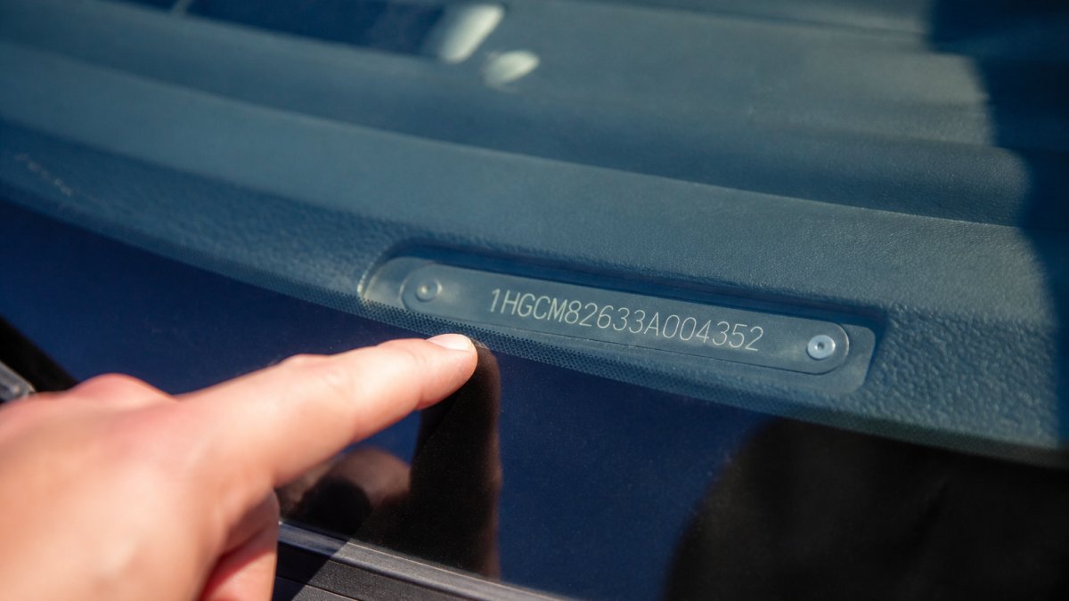 Close-up of a vehicle identification number (VIN) plate mounted on a car's dashboard, with a person's finger pointing to the 17-character alphanumeric code under natural sunlight