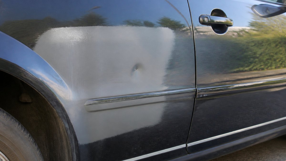 Close-up of a used car's side panel showing mismatched paint color and a small dent from previous damage, with natural daylight highlighting the repair work