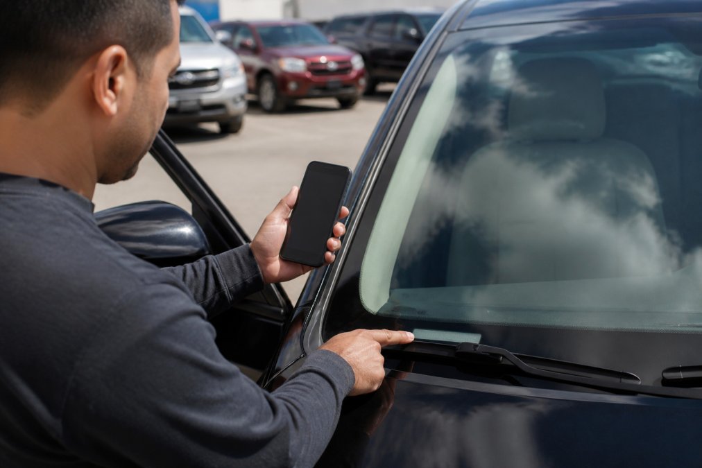 A person holding a smartphone displaying a VIN number while standing beside an open car door, checking the vehicle identification plate visible through the windshield, daylight reflecting off the glass