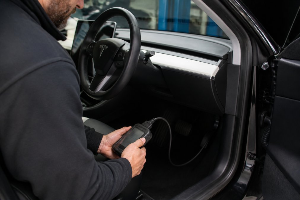 Professional technician holding a diagnostic scanner connected to an electric vehicle's on-board diagnostics port in a well-lit workshop, with the vehicle's dashboard visible in the background