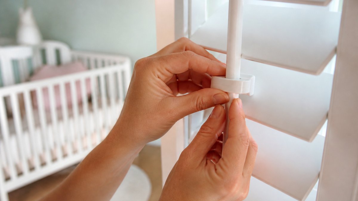 Close-up of an adult's hands fitting a small child safety clip to the tilt rod of a white plantation shutter in a bright nursery, with a cot visible softly out of focus in the background