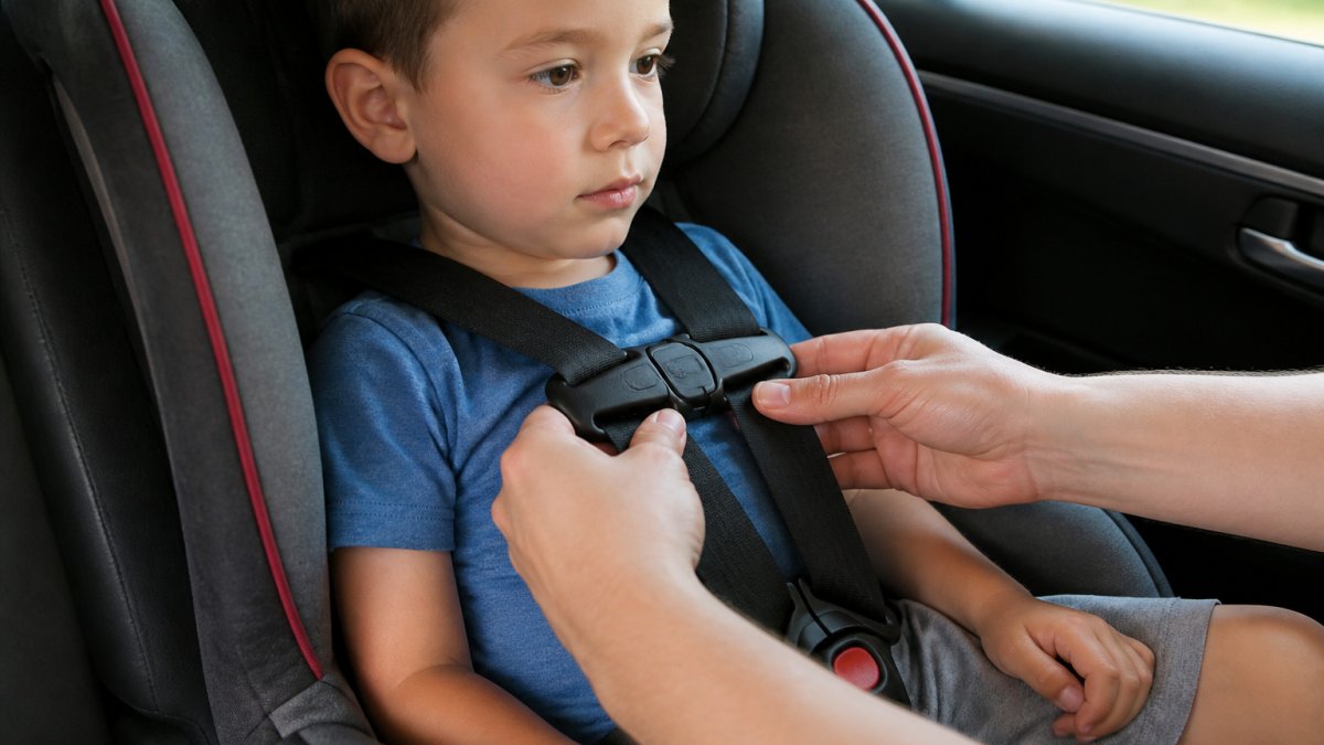A parent's hands doing the pinch test on a child's car seat harness straps over the chest clip, child seated in a forward-facing seat in a parked car