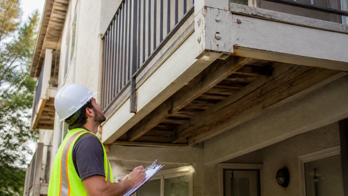 A licensed inspector in a hard hat and safety vest examining the underside of an elevated exterior balcony on a multi-story residential building, clipboard in hand, daylight, close view of railing and deck connections