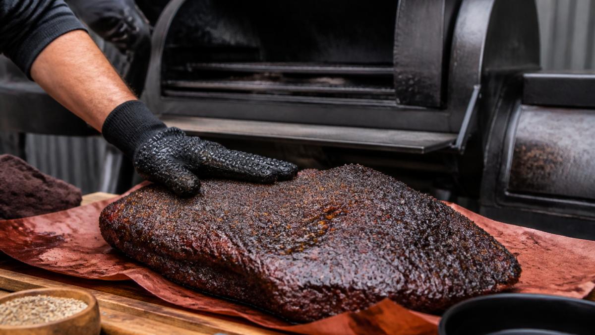 Close-up of a whole smoked beef brisket resting on butcher paper, dark mahogany bark visible, a hand in a heat-proof glove pressing the surface, offset smoker door open in the background