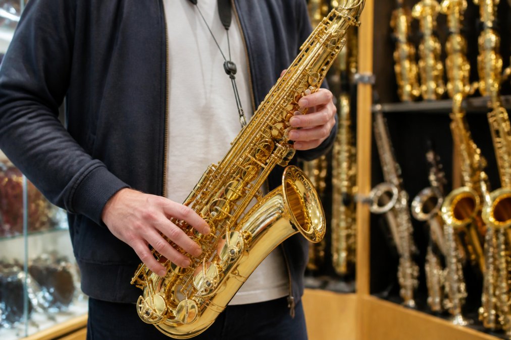 A close-up of a person's hands holding a shiny alto saxophone in a music shop, with other instruments blurred in the background