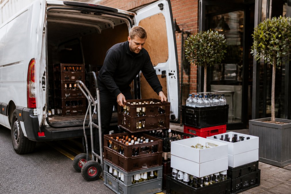 A delivery driver carefully unloading crates of premium wines and spirits from a refrigerated van at a hospitality venue's entrance, showing professional handling and organised stock arrangement
