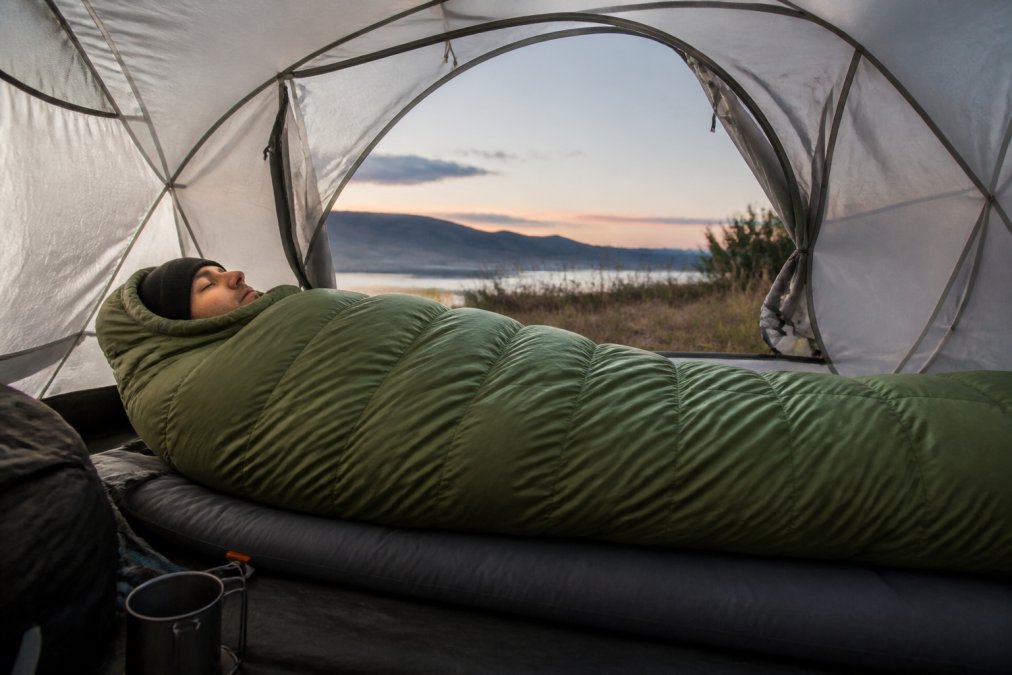 Camper lying in a mummy-shaped sleeping bag inside a tent during evening light, showing how the bag contours around the body