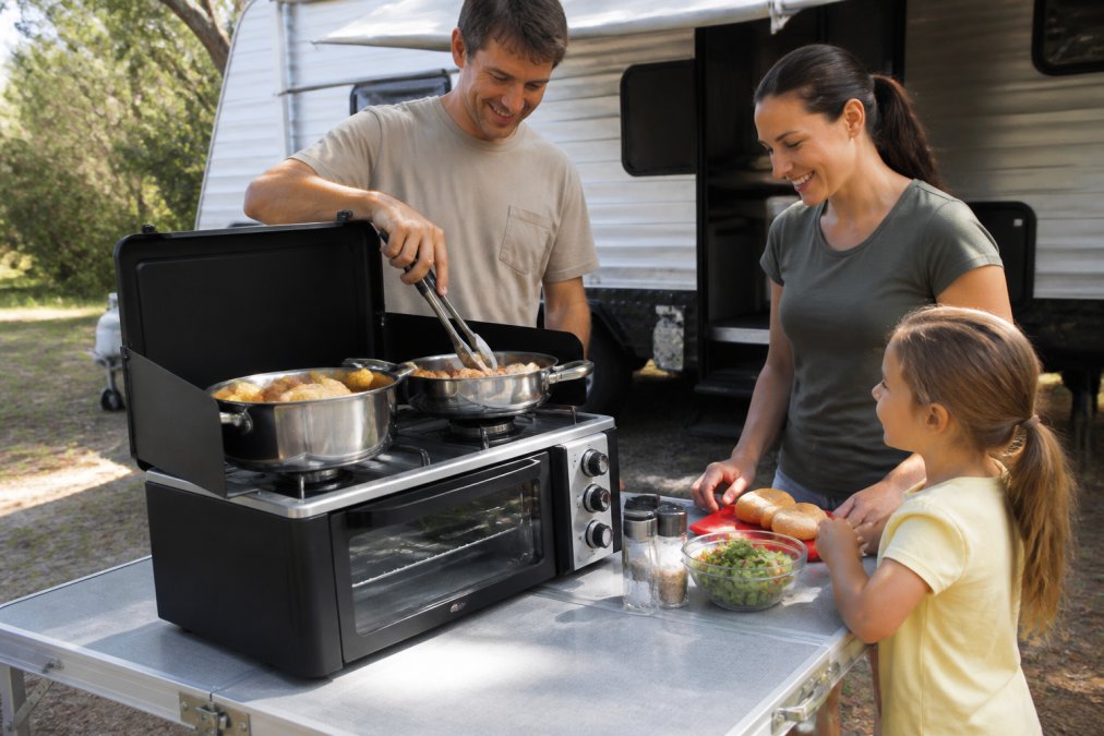 A family cooking a full meal on a two-burner portable gas camping stove with oven at a campsite table during the day, with a caravan in the background