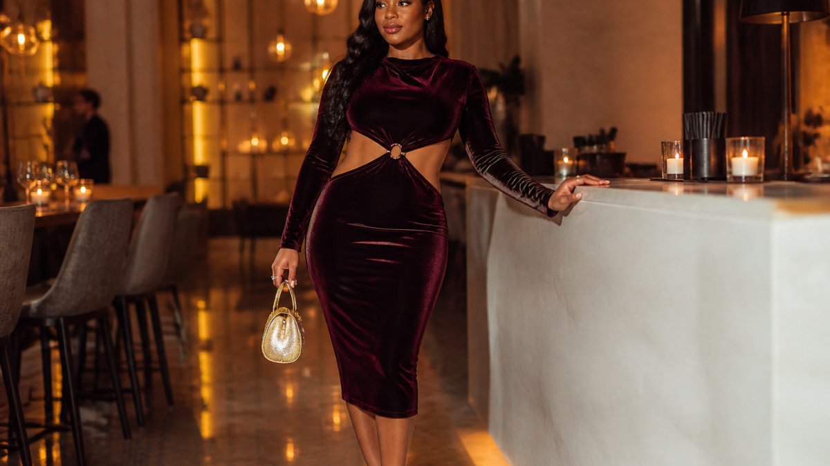 Woman wearing a midi-length velvet cocktail dress with long sleeves and cutout details, posing confidently at an upscale event venue with warm ambient lighting
