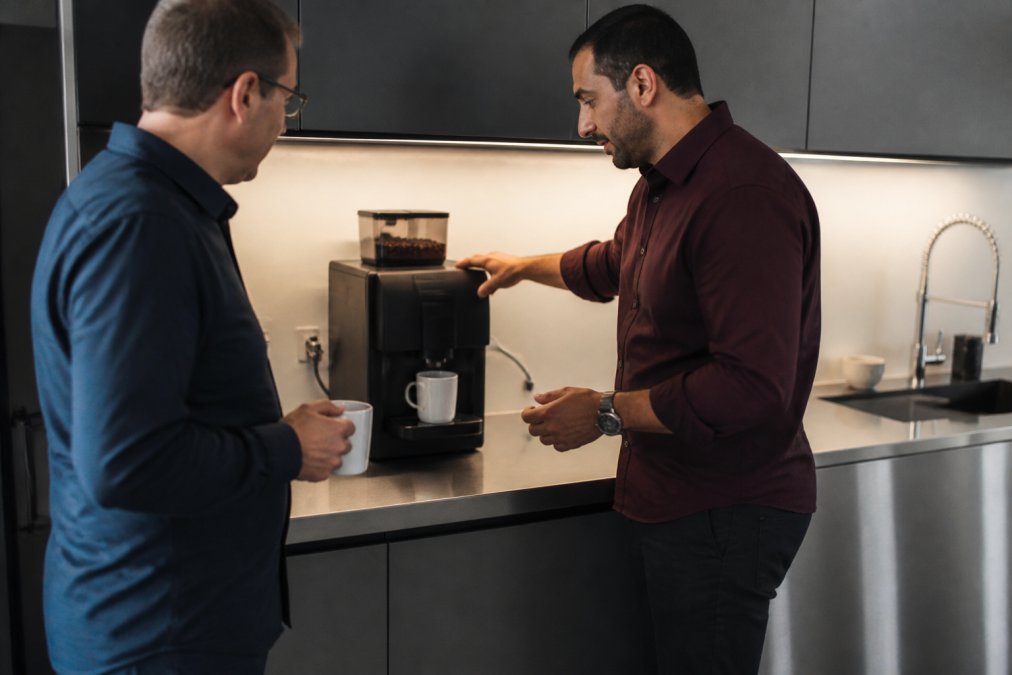 A compact bean-to-cup coffee machine on a stainless steel countertop in a small, tidy office kitchen, with a water line connected and a mug waiting underneath