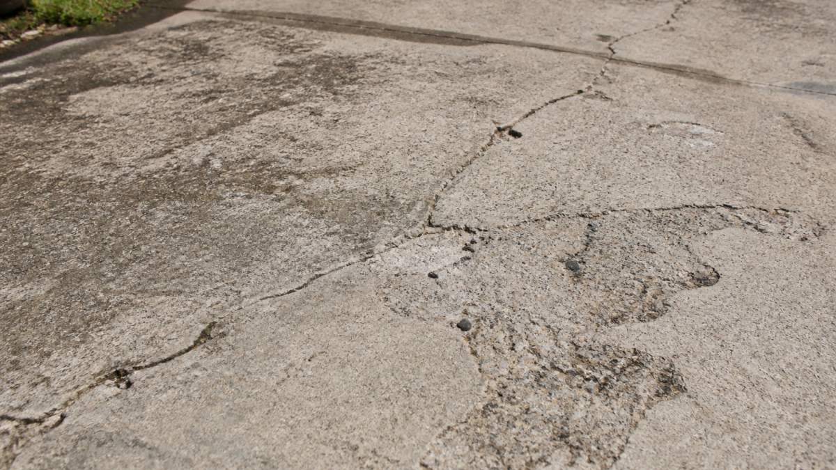 Close-up of deteriorating concrete driveway showing visible cracks, stains, and surface spalling with natural daylight highlighting damage