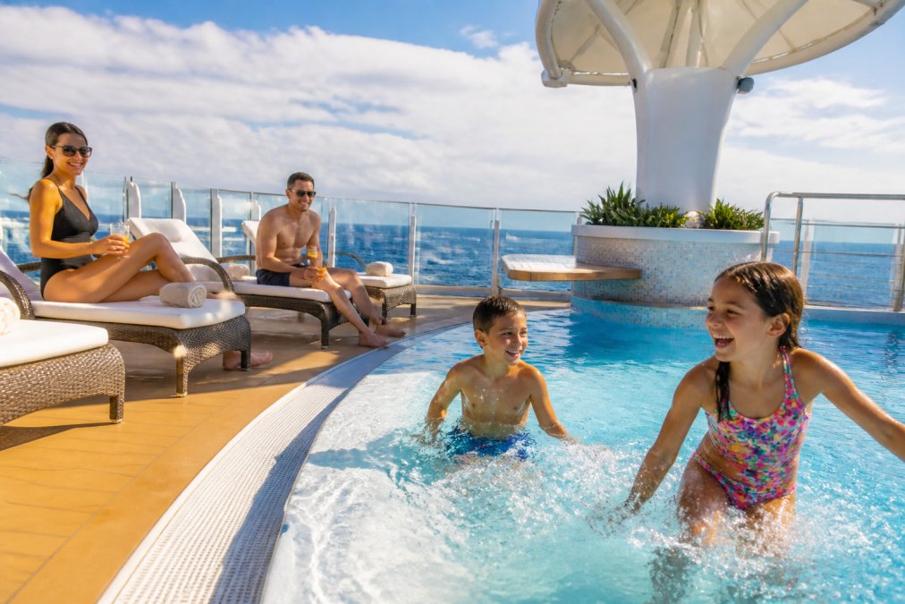 Family of four enjoying activities on a cruise ship deck, with children playing in a pool area and parents relaxing nearby, ocean and sky visible in background