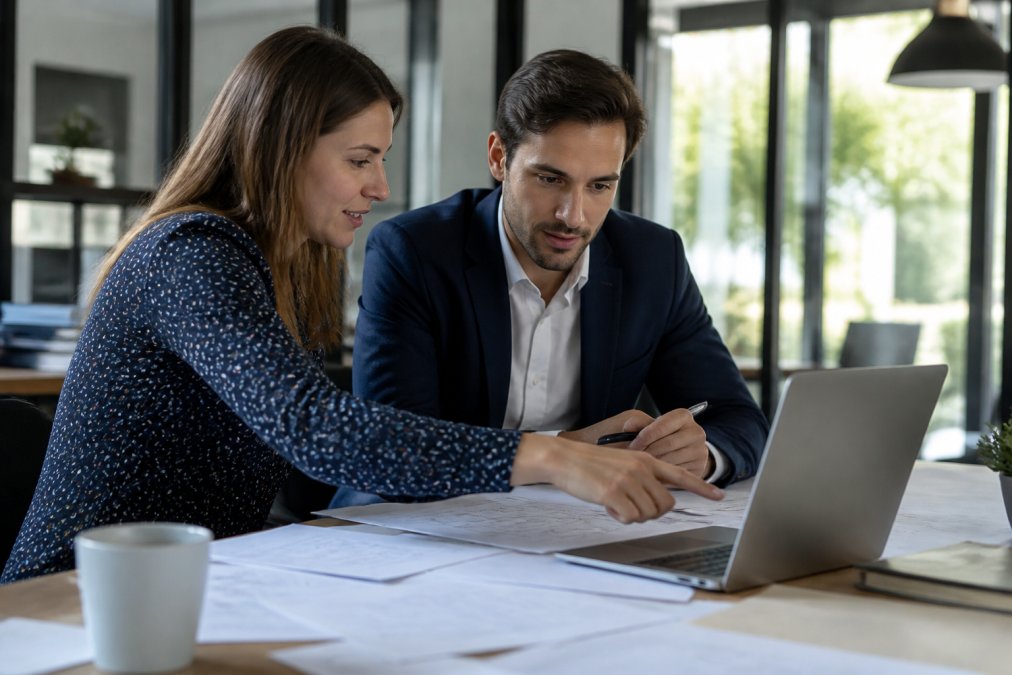Two professionals in a modern office setting having a discussion over a laptop, one pointing at the screen while reviewing project requirements and documentation on papers spread across the desk, natural daylight from windows