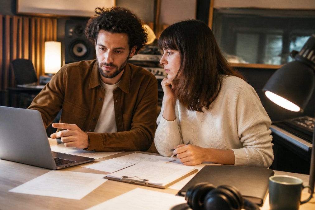 Music producer and label manager reviewing artist contracts and sublabel structure on a laptop in a home studio environment with warm lighting from desk lamp