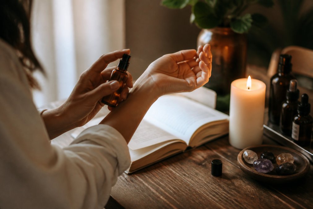 A person's hands applying fragrance from a small amber glass bottle to their wrist, with a lit candle and an open book on a wooden table in warm, soft light
