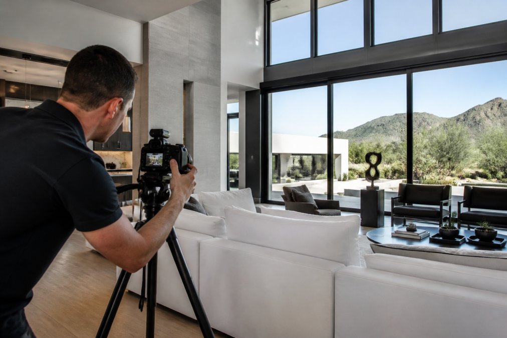 A professional photographer adjusting a camera on a tripod in the bright, spacious living room of a high-end modern home with large windows and desert views