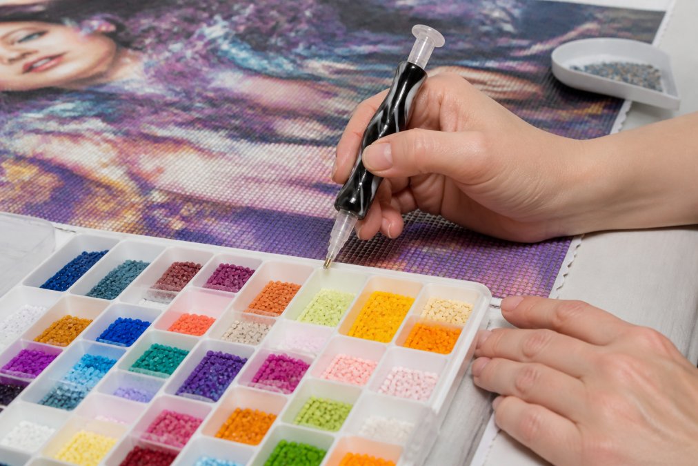 Close-up of hands holding ergonomic diamond drill pens with comfort-grip design and specialized tray showing organized diamond placement by color during active diamond painting session, natural studio lighting
