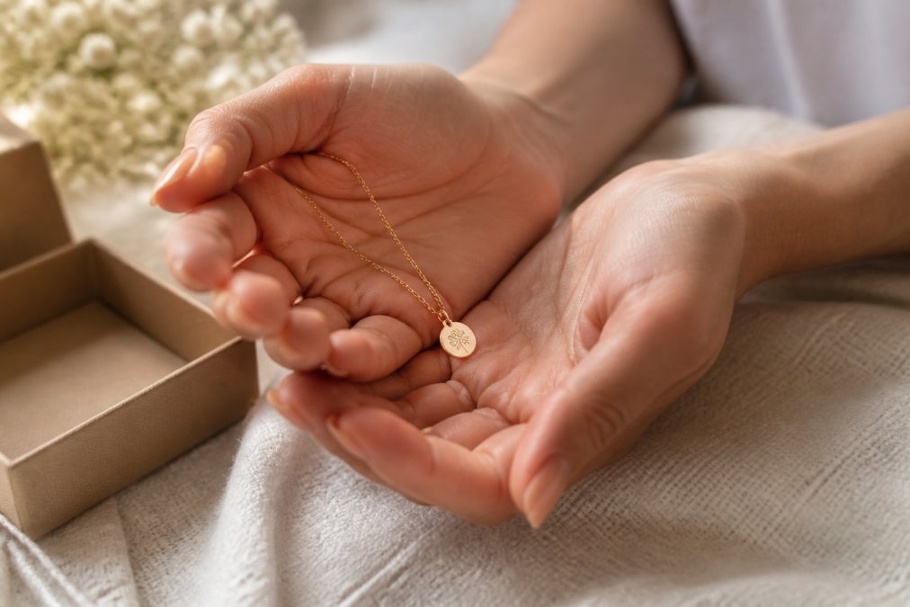 Close-up of a woman's hands holding a delicate personalized necklace with a small engraved charm, resting on a soft fabric surface in warm natural light
