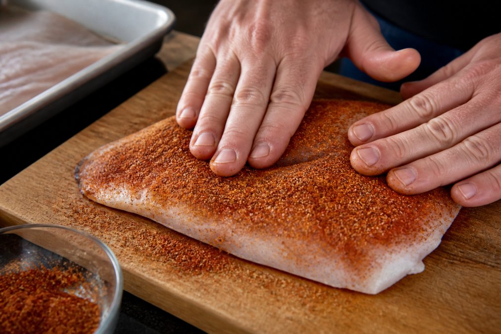 Close-up of hands massaging a dry rub onto a fresh fish fillet on a cutting board, with the rub forming a visible crust on the surface