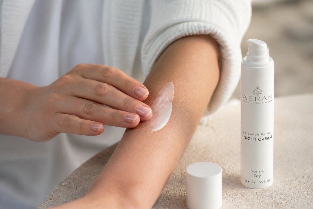 Close-up of hands applying a rich moisturizer cream to dry forearm skin, showing the texture and absorption, with warm natural lighting highlighting the skin's texture