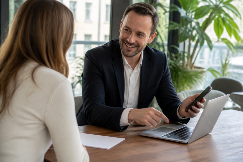A real estate professional using a laptop and smartphone to show property listings and market data to a client in a bright, modern office with natural lighting