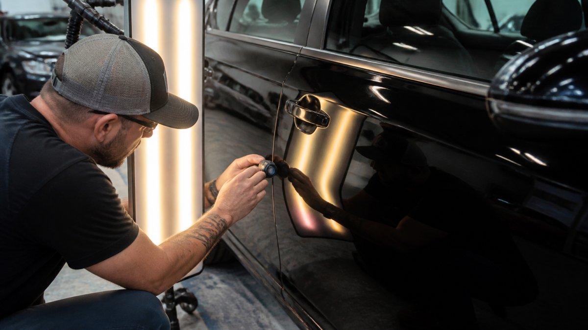 Professional PDR technician inspecting a vehicle's dented door panel with specialized lighting tools and magnification, demonstrating hands-on inspection technique in a well-equipped repair facility