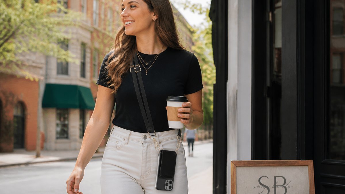 A woman in a casual outfit walking through a city street with a crossbody phone case hanging at her hip, her hands free while she holds a coffee cup, natural daylight