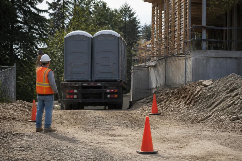 A portable restroom delivery truck backing carefully into a tight gravel construction site entrance, with a worker in a safety vest guiding the driver and orange cones marking the path