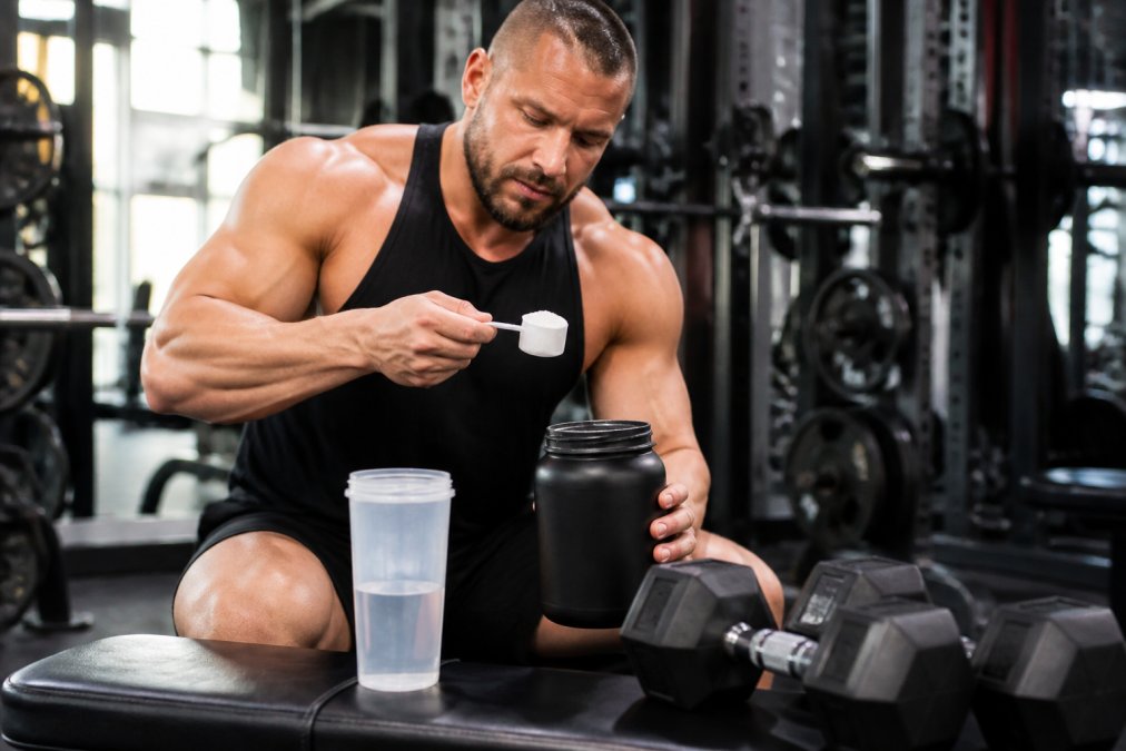 Athlete in gym setting preparing to take a scoop of pre-workout powder, with water bottle and dumbbells visible on the bench, under bright gym lighting