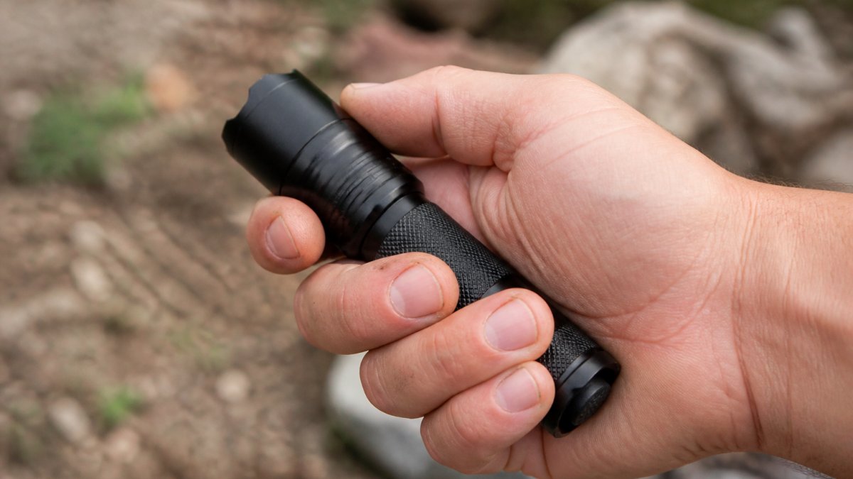 Close-up of a rugged EDC flashlight being gripped firmly in a hand outdoors, showing textured aluminum body, knurled grip, and solid metal construction in natural daylight