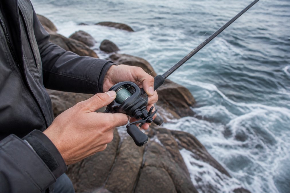 Close-up of an angler's hands holding a baitcasting reel loaded with braided line, standing on a rocky shoreline with ocean water in the background